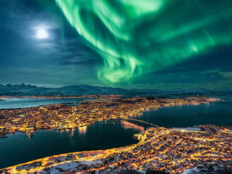 Aurora Borealis dancing over the city of Tromso and full moon.  Tromso Urban Skyline and winter night landscape with Northern Lights (Aurora Polaris) on a cold night, with starry sky, polar lights and snowcapped mountains on Kvaloya Island in the background. Cityscape with Brightly Street Light of the Arctic Capital, Northern Norway